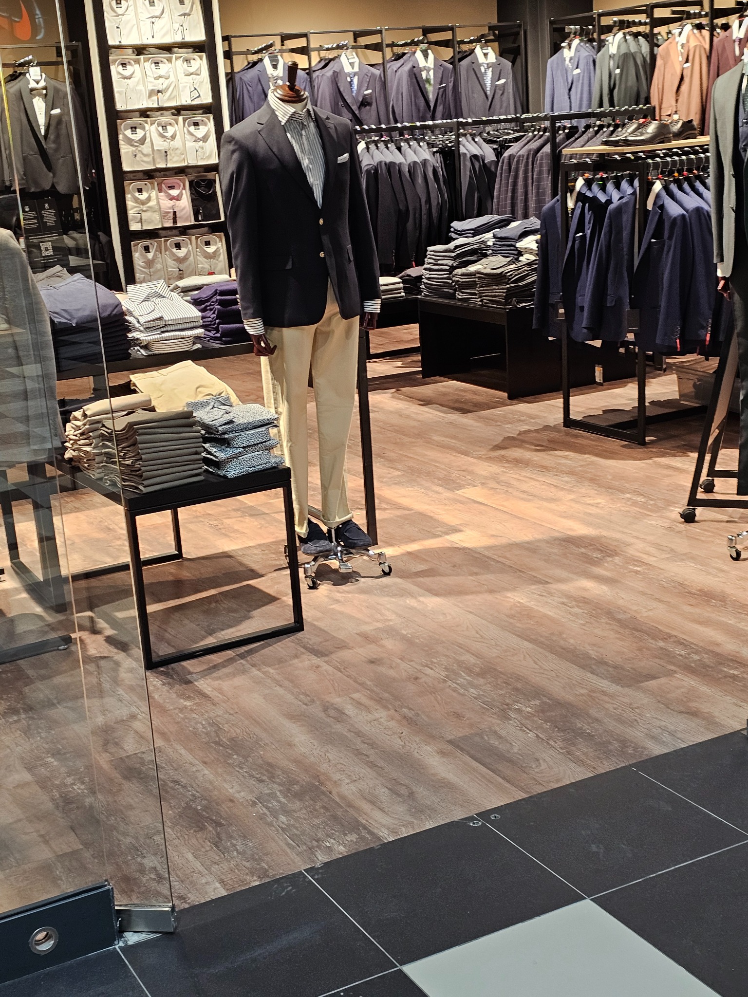 Wnętrze sklepu odzieżowego z drewnopodobną podłogą, prezentacja męskich garniturów, koszul i spodni na manekinach i wieszakach.