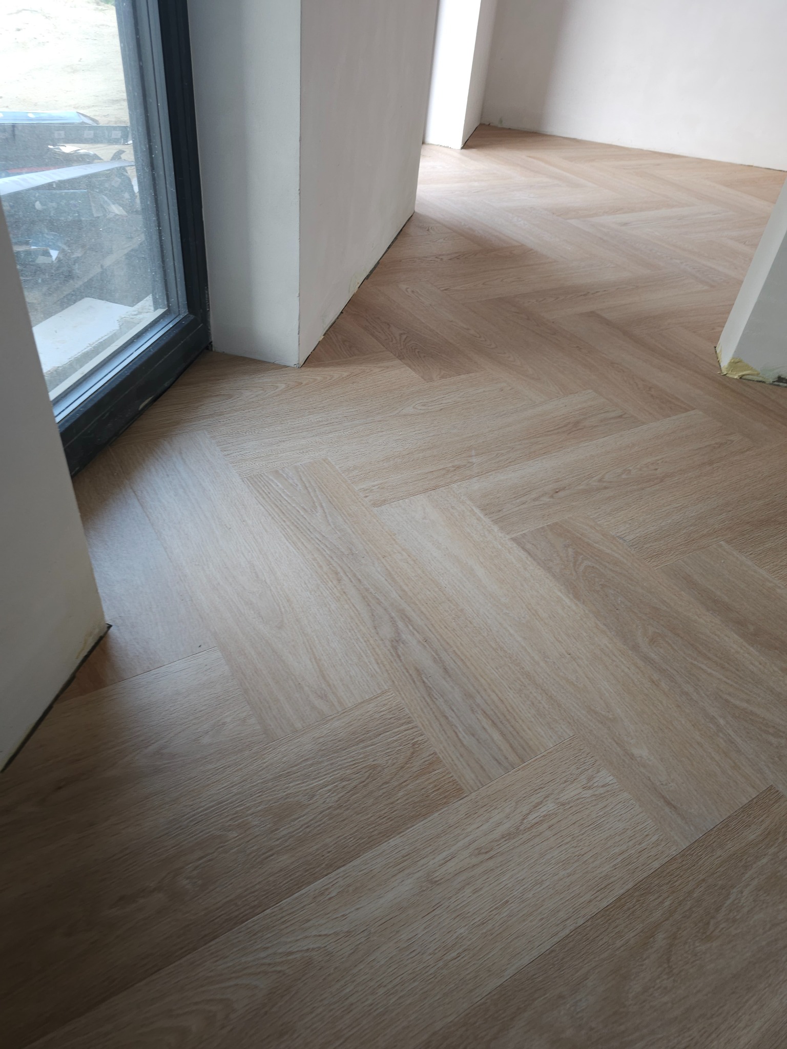 Jasne panele ułożone w jodełkę, widok z góry, fragment okna i ściany w tle, wykończenie w trakcie prac.