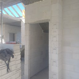 Topolowa 2C - Wnętrze budynku w trakcie budowy z białych bloczków, widoczny otwór drzwiowy z charakterystycznym wykończeniem narożników, puszka z pianką montażową na ułożonych bloczkach i torba budowlana w rogu...