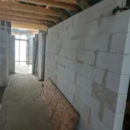 Wnętrze budynku w trakcie budowy, widoczne ściany z białych bloczków, drewniana konstrukcja dachu i podłoga w stanie surowym, z deską opartą o ścianę.