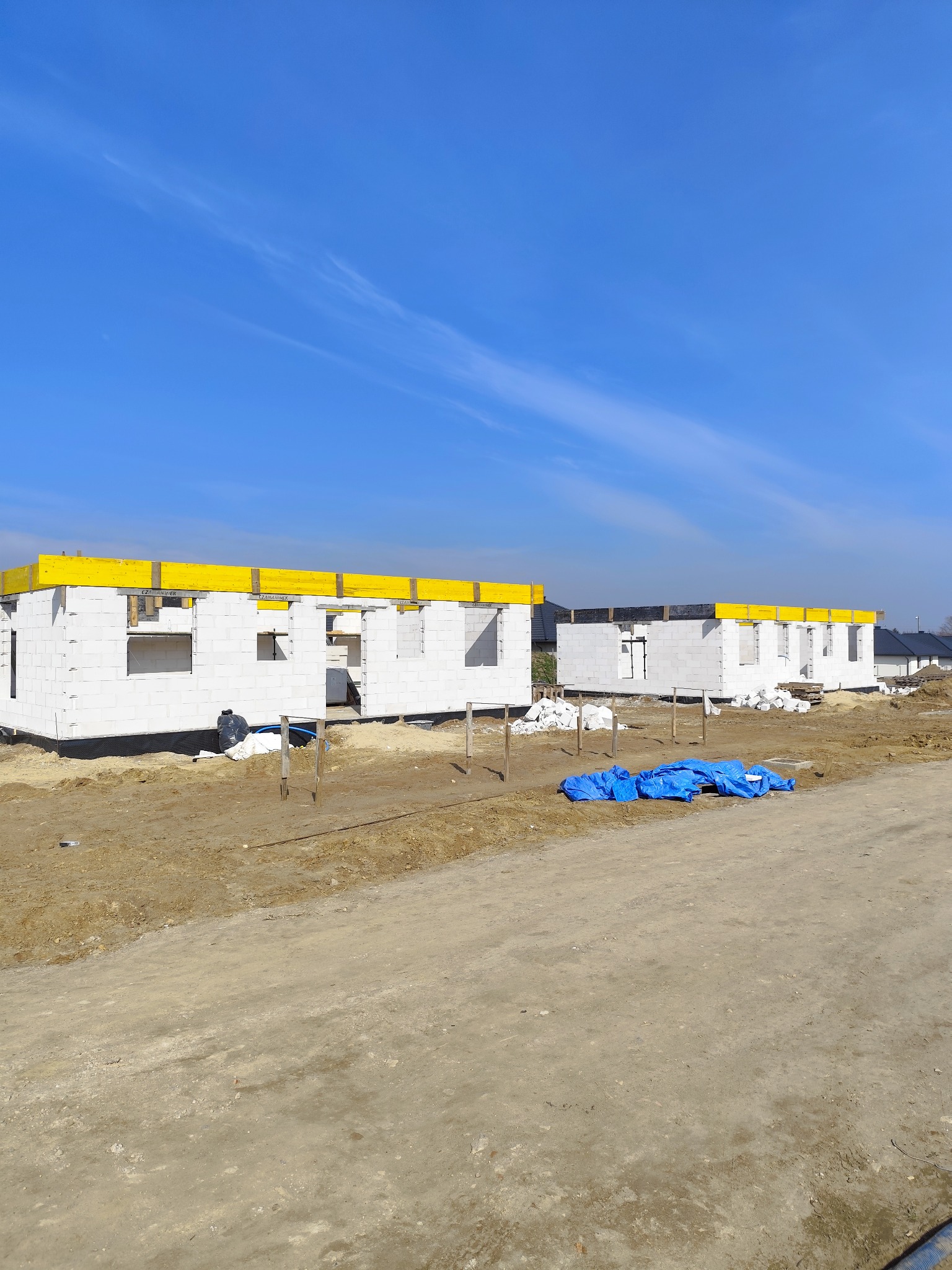 Wznoszenie ścian parterowych domów z białych bloczków, z żółtymi elementami szalunkowymi na stropie i niebieską plandeką w tle, na placu budowy w słoneczny dzień.