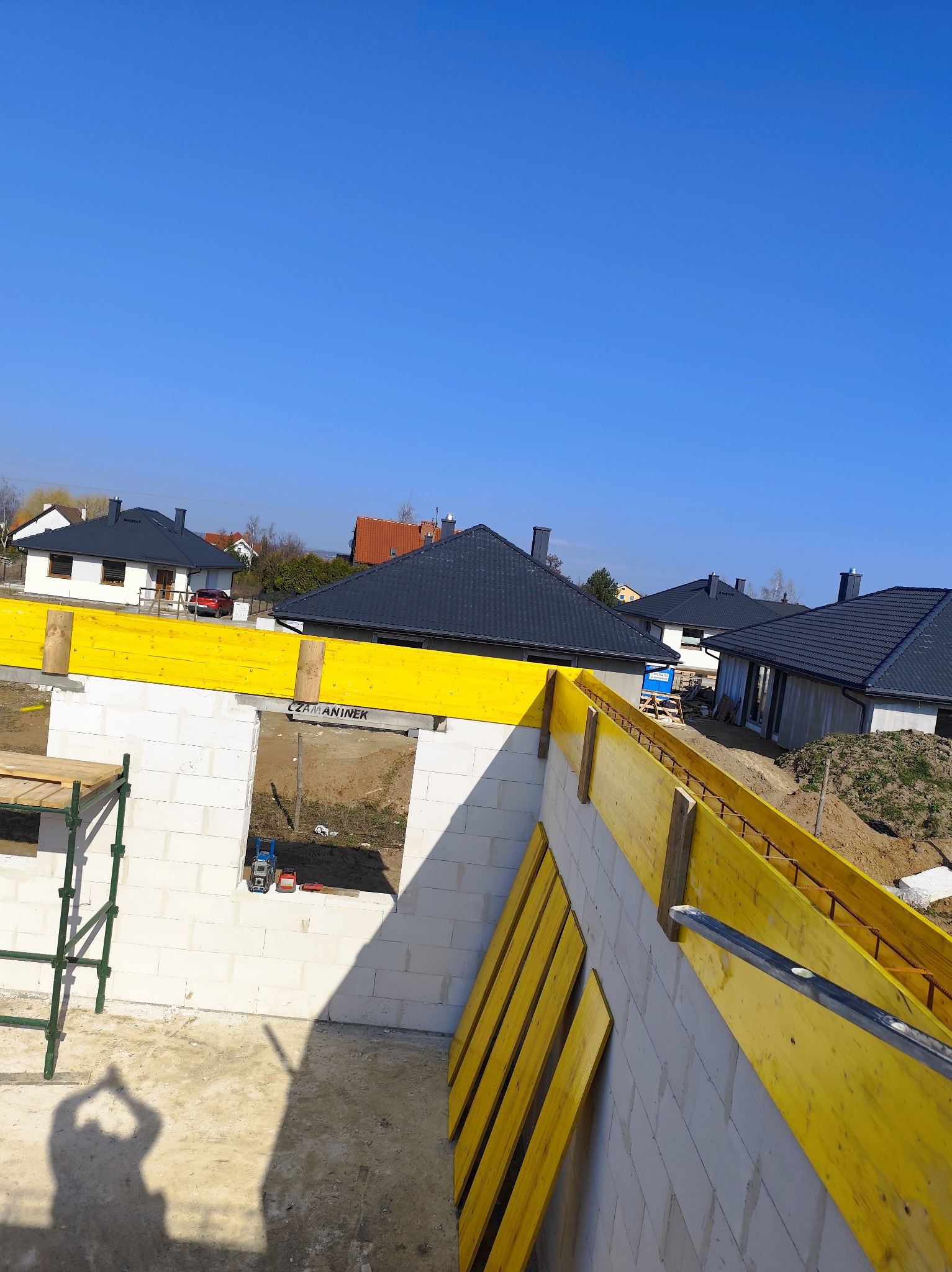 Wznoszenie ścian z bloczków betonu komórkowego Ytong, z widocznym żółtym szalunkiem i zbrojeniami, w tle zabudowa jednorodzinna i błękitne niebo, cień osoby fotografującej w dolnej części kadru.