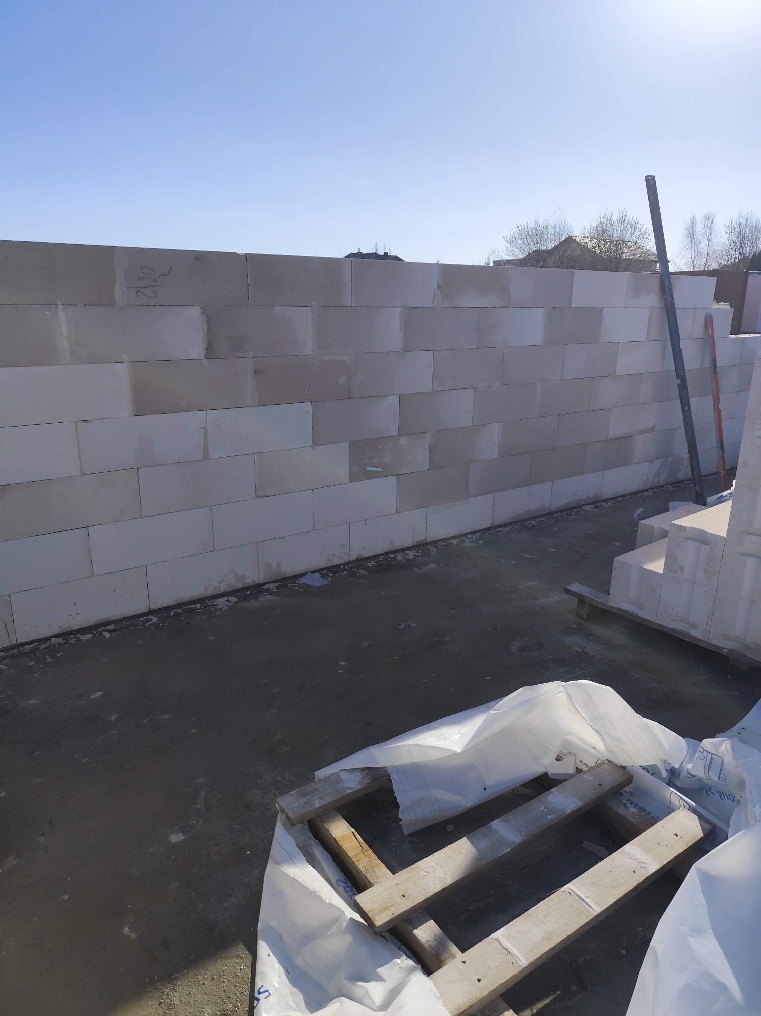 Wzniesiona ściana z białych bloczków betonowych na placu budowy, widoczne narzędzia murarskie i paleta z materiałami budowlanymi pod folią, jasne niebo w tle.