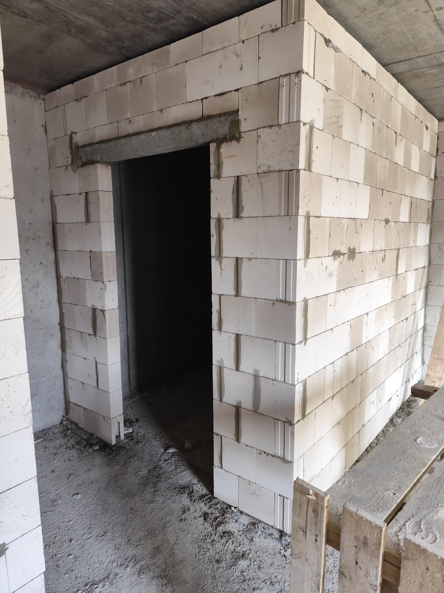 Wnętrze budynku w trakcie budowy z białych bloczków, widoczne nadproże nad otworem drzwiowym, surowy betonowy strop i paleta drewniana w rogu pomieszczenia.