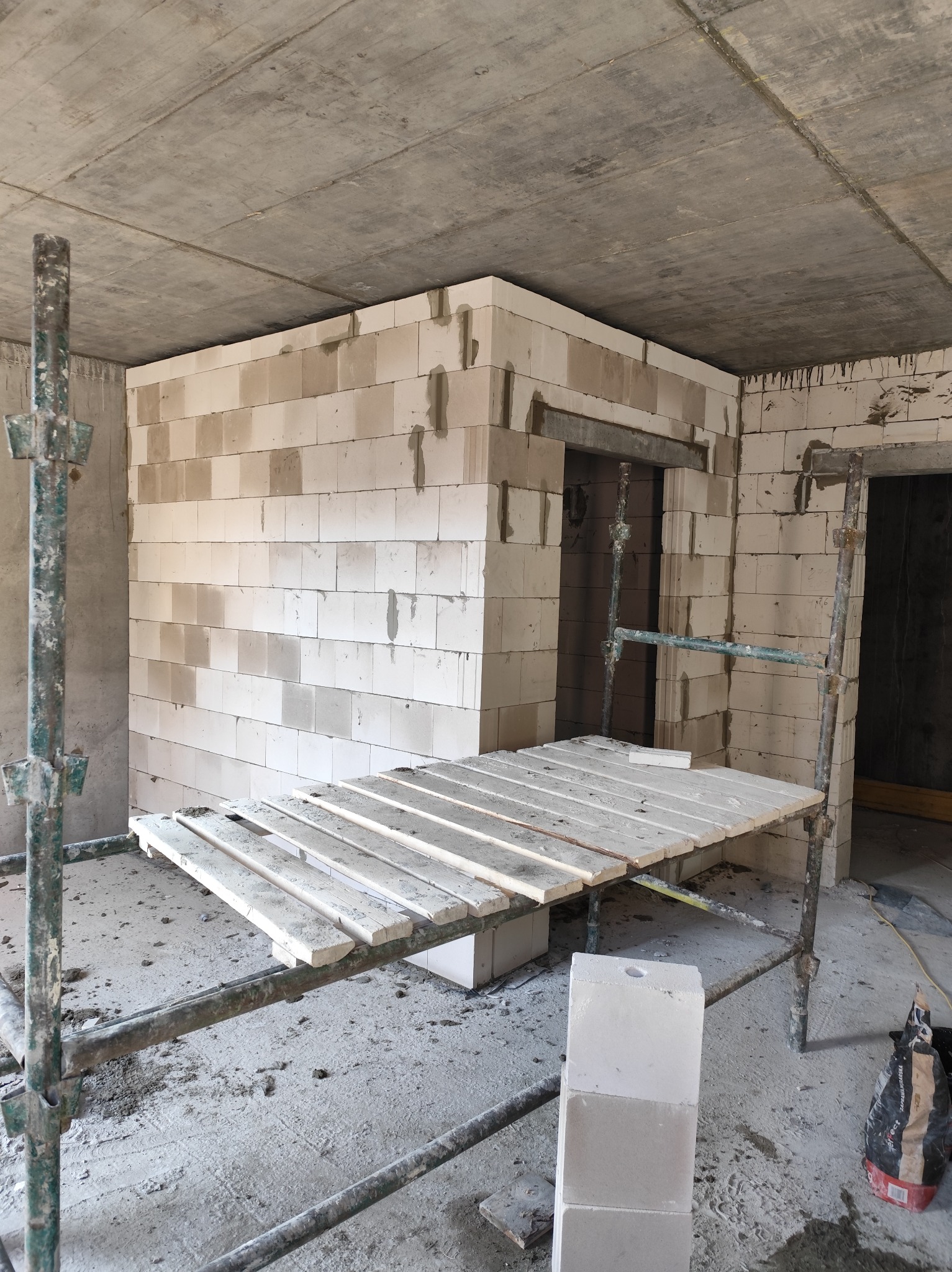 Wnętrze w trakcie budowy w Miłobądzu: widok na wznoszone ściany z białych bloczków, częściowo osadzone nadproża drzwiowe, z widocznym rusztowaniem i workiem zaprawy na podłodze.