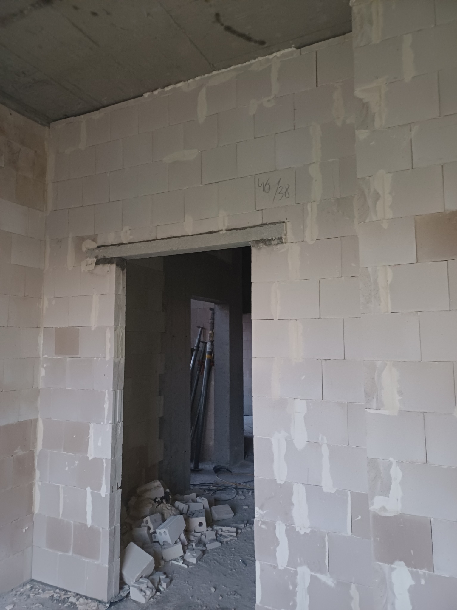 Wnętrze w trakcie budowy z widoczną ścianą z białych bloczków, nadprożem nad otworem drzwiowym i zapisanym numerem '46/38' na jednej z cegieł.