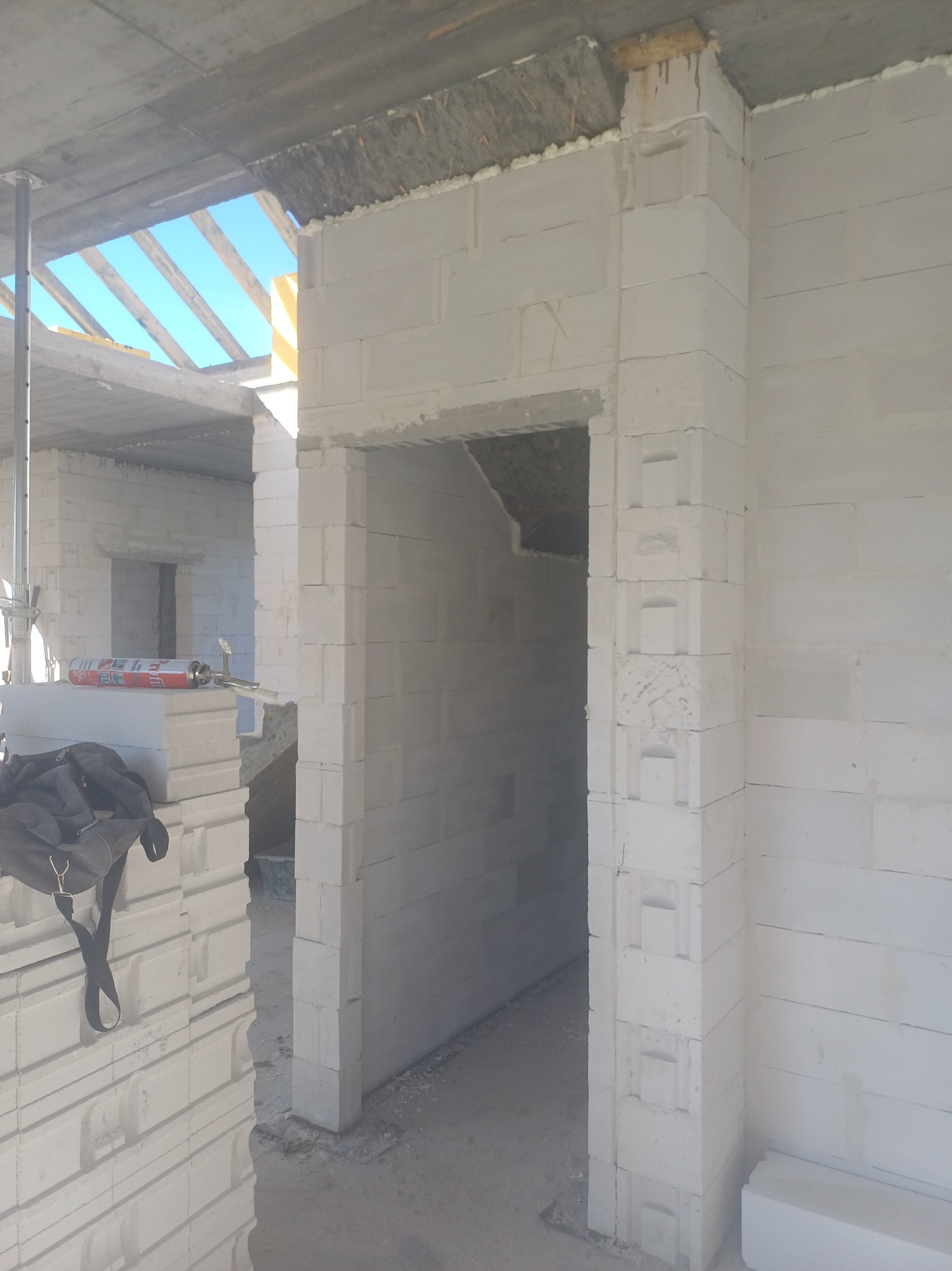 Wnętrze budynku w trakcie budowy z białych bloczków, widoczny otwór drzwiowy z charakterystycznym wykończeniem narożników, puszka z pianką montażową na ułożonych bloczkach i torba budowlana w rogu...