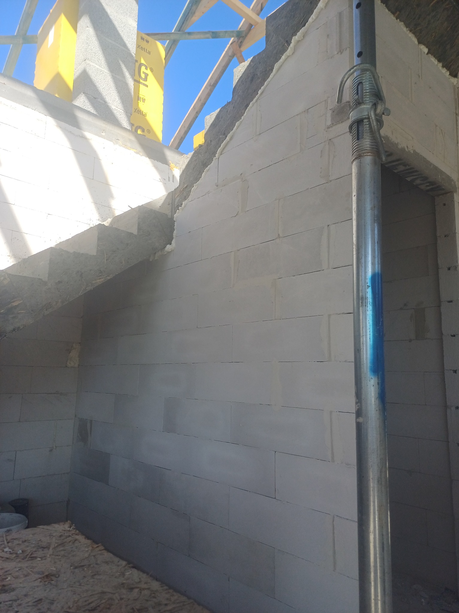 Wnętrze budynku w trakcie budowy, ściany z białych bloczków, widoczna metalowa podpórka i fragment drewnianej konstrukcji dachu.