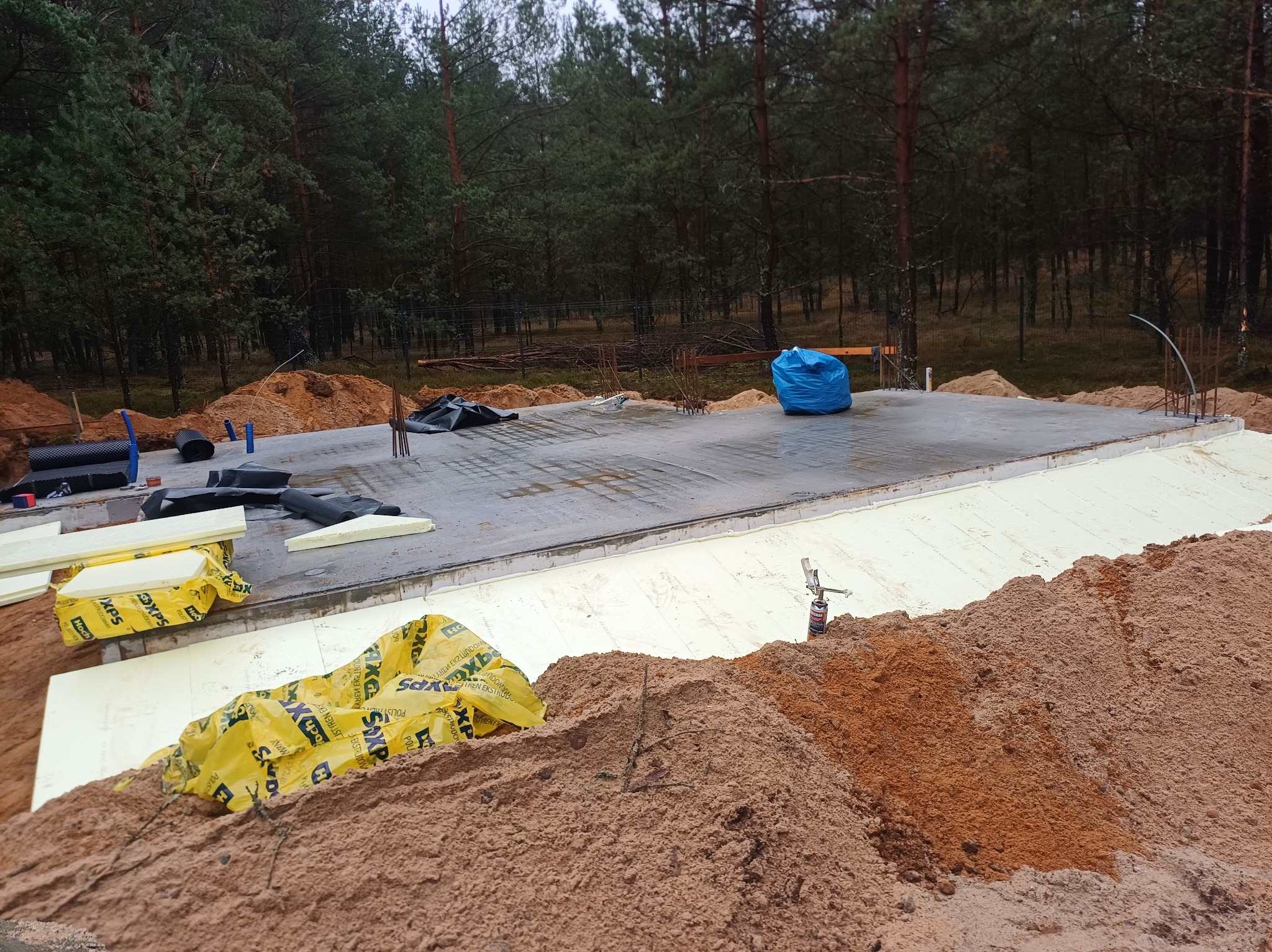 Przygotowany fundament pod budynek, ocieplony styropianem XPS, z widocznymi elementami zbrojenia i instalacji, na tle zalesionego terenu.
