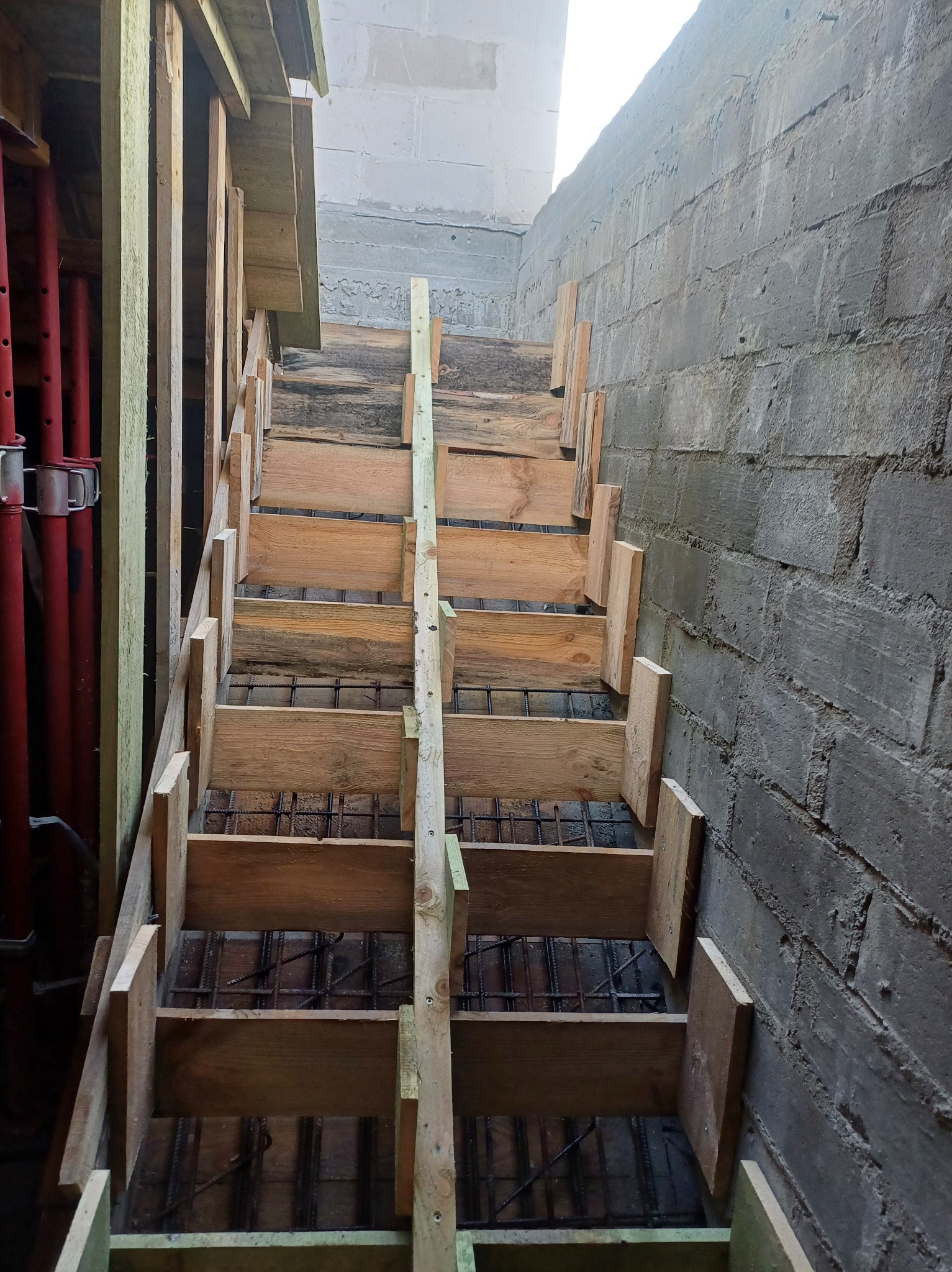 Drewniane szalunki schodów z widocznym zbrojeniem i fragmentem murowanej ściany z bloczków betonowych. Czerwone podpory szalunkowe widoczne z lewej strony.