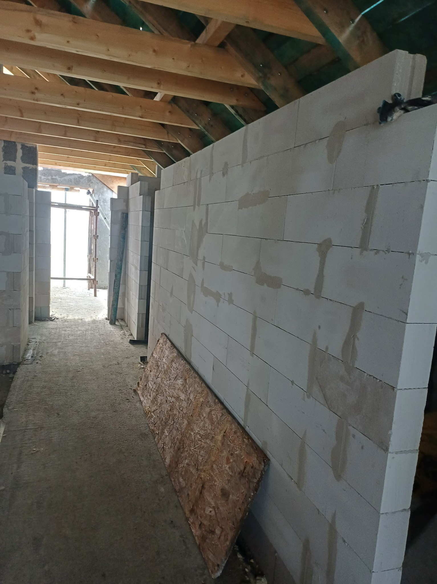 Wnętrze budynku w trakcie budowy, widoczne ściany z białych bloczków, drewniana konstrukcja dachu i podłoga w stanie surowym, z deską opartą o ścianę.