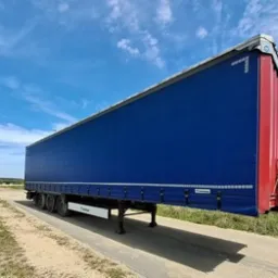 Niebieska naczepa TIR-a marki Krone z czerwonym tyłem, zaparkowana na betonowej nawierzchni, widok z boku pod lekkim kątem, błękitne niebo w tle.