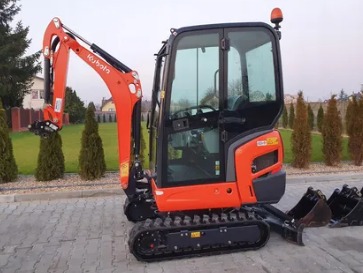 Pomarańczowa minikoparka Kubota z kabiną, stojąca na zewnątrz z kilkoma łyżkami z boku, widok z boku.