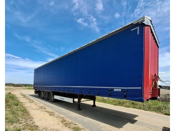 Niebieska naczepa TIR-a marki Krone z czerwonym tyłem, zaparkowana na betonowej nawierzchni, widok z boku pod lekkim kątem, błękitne niebo w tle.