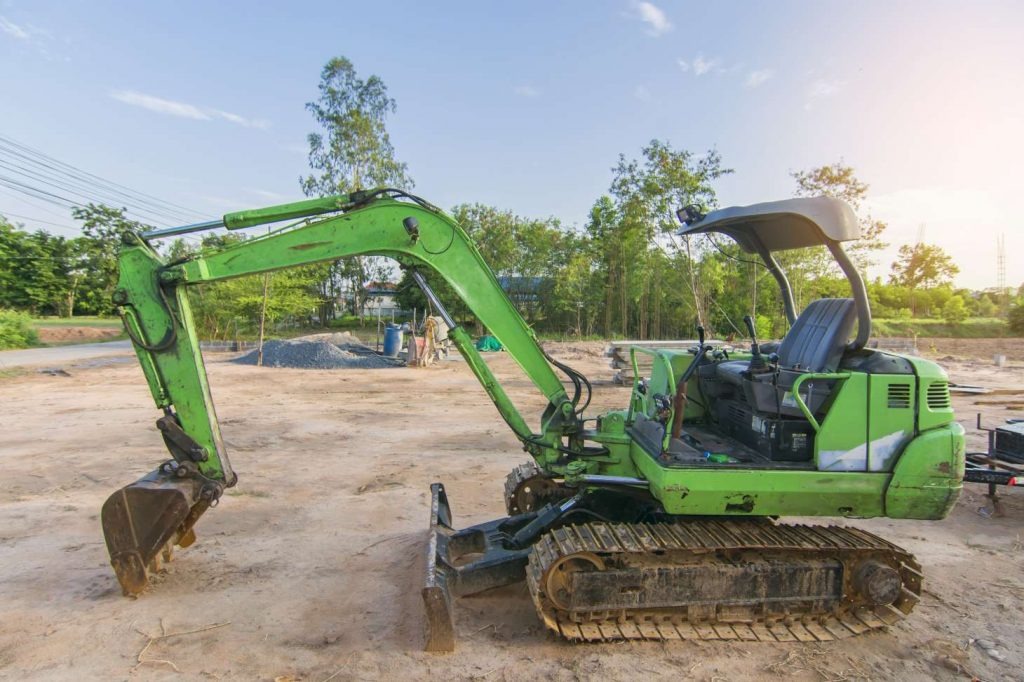 Używana, zielona mini koparka na gąsienicach w Olsztynie, zaparkowana na placu budowy, w tle drzewa i błękitne niebo.