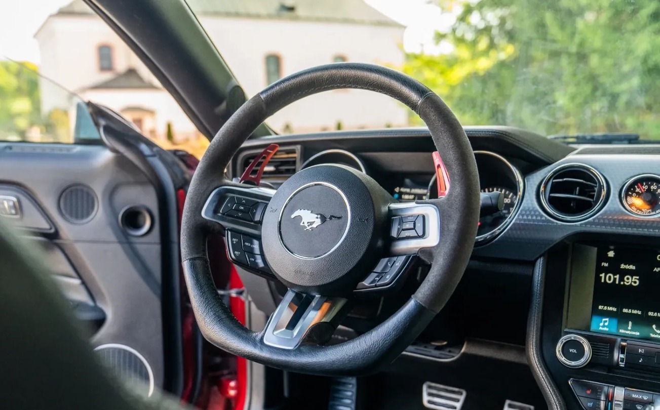 Wnętrze samochodu sportowego: kierownica z logo konia, czerwone elementy, deska rozdzielcza z wyświetlaczem, widok na budynek w tle.