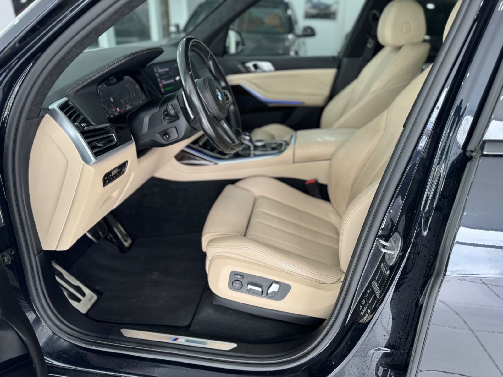 Wnętrze czarnego samochodu z beżową skórzaną tapicerką, widok z otwartych drzwi na przedni fotel pasażera i deskę rozdzielczą, z detalami takimi jak aluminiowe pedały i logo M Performance.