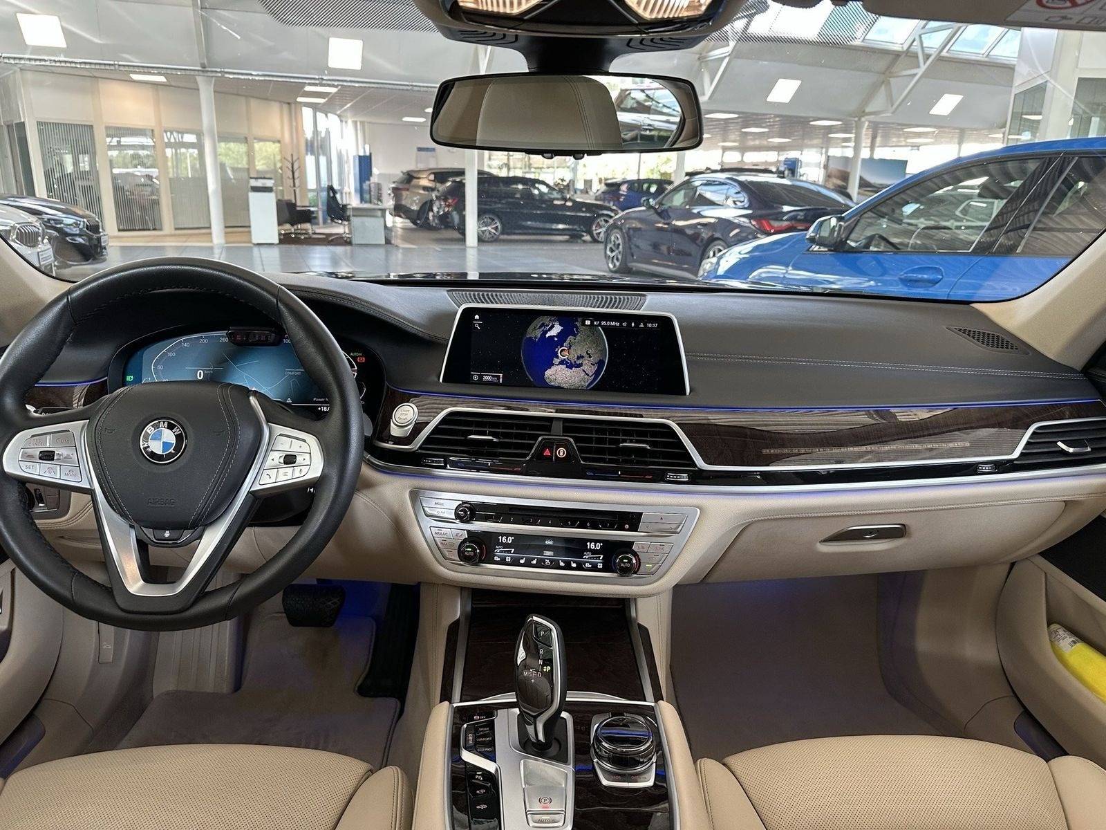 Wnętrze luksusowego samochodu BMW z beżową skórzaną tapicerką, widok na deskę rozdzielczą z wyświetlaczem pokazującym mapę świata, kierownicę z logo BMW oraz automatyczną skrzynię biegów. W tle...