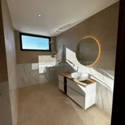 Nowoczesna łazienka z kabiną prysznicową typu walk-in, umywalką nablatową i okrągłym lustrem z podświetleniem LED. Beżowe płytki i marmurowe akcenty.