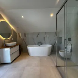 Nowoczesna łazienka na poddaszu z wolnostojącą wanną, kabiną prysznicową ze szklanymi drzwiami i okrągłym lustrem z podświetleniem LED. Minimalistyczny design.