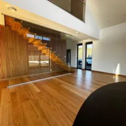 Nowoczesne schody drewniane z przeszkloną balustradą w jasnym wnętrzu domu. Parkiet z naturalnego drewna, minimalistyczny design, duże okna w tle.