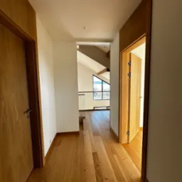 Korytarz z drewnianą podłogą i drzwiami z tego samego materiału. W tle widoczne okno z widokiem na krajobraz. Minimalistyczny design, naturalne światło.