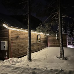 INWESTNOR SPÓŁKA Z OGRANICZONĄ ODPOWIEDZIALNOŚCIĄ - Oświetlona drewniana sauna w zimowej scenerii nocą. Śnieg pokrywa teren, a drzewa iglaste dodają uroku. Widoczna szopa na drewno i dekoracyjne lampki na dachu.