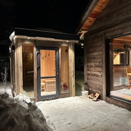 INWESTNOR SPÓŁKA Z OGRANICZONĄ ODPOWIEDZIALNOŚCIĄ - Drewniana sauna zewnętrzna z przeszklonymi ścianami i drzwiami, oświetlona, w zimowej scenerii z pokrywą śnieżną. Widoczny fragment drewnianego domu obok.