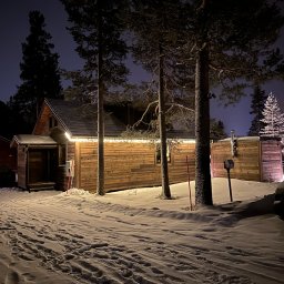 INWESTNOR SPÓŁKA Z OGRANICZONĄ ODPOWIEDZIALNOŚCIĄ - Zimowy krajobraz z drewnianą sauną podświetloną girlandą światełek. Śnieg pokrywa ziemię, a wysokie sosny dodają uroku nocnej scenerii.