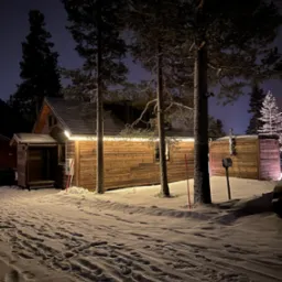 Zimowy krajobraz z drewnianą sauną podświetloną girlandą światełek. Śnieg pokrywa ziemię, a wysokie sosny dodają uroku nocnej scenerii.