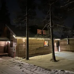 Oświetlony, drewniany dom w zimowej scenerii, widoczny nocą, z dekoracyjnymi lampkami i śniegiem. Charakterystyczna architektura i oświetlenie.