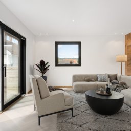 INWESTNOR SPÓŁKA Z OGRANICZONĄ ODPOWIEDZIALNOŚCIĄ - Jasny salon z drewnianą ścianą i wyjściem na taras przez przesuwne drzwi balkonowe. W tle widoczny dom z białymi ścianami i czerwonym dachem.