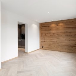 INWESTNOR SPÓŁKA Z OGRANICZONĄ ODPOWIEDZIALNOŚCIĄ - Jasny parkiet ułożony w jodełkę francuską w przestronnym wnętrzu z drewnianą ścianą akcentującą i widokiem na kuchnię. Minimalistyczny design.