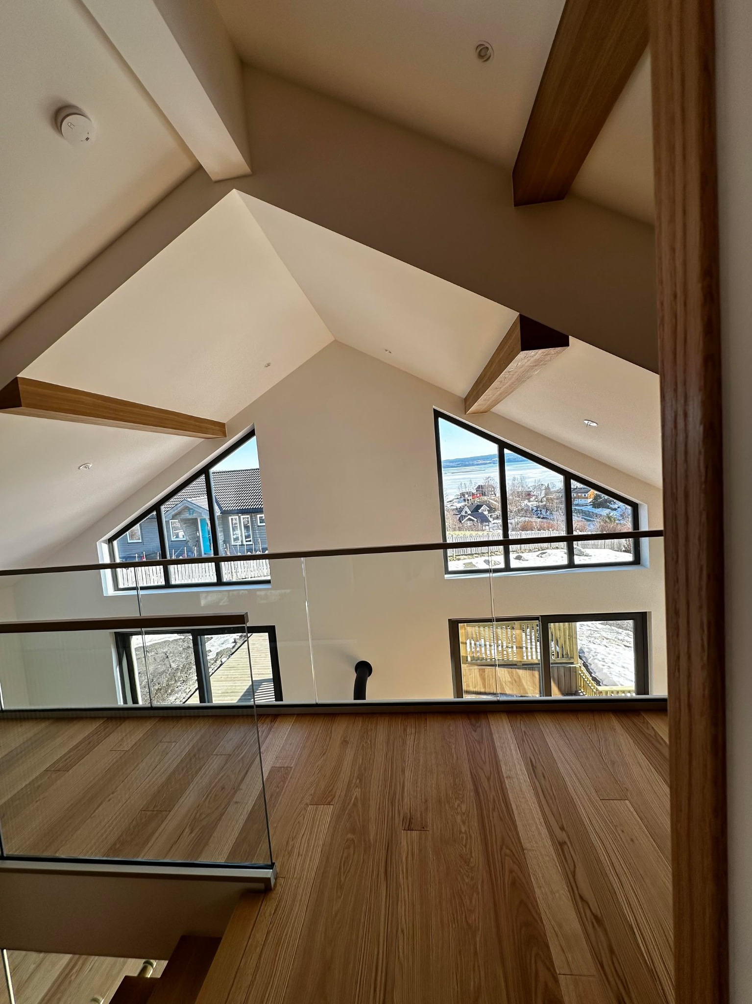 Nowoczesne wnętrze domu z drewnianą podłogą i oknami trójkątnymi z widokiem na zimowy krajobraz. Szklana balustrada, drewniane belki stropowe.