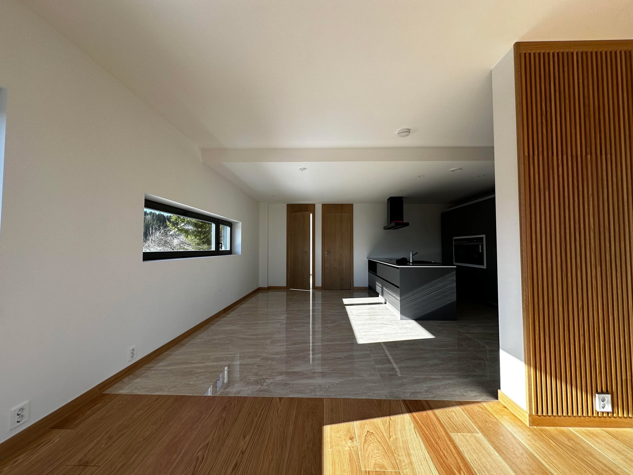 Jasne wnętrze domu z drewnianą podłogą przechodzącą w marmurowe płytki, drewniane drzwi i dekoracyjna ściana z drewnianych listew. Nowoczesna kuchnia w tle.