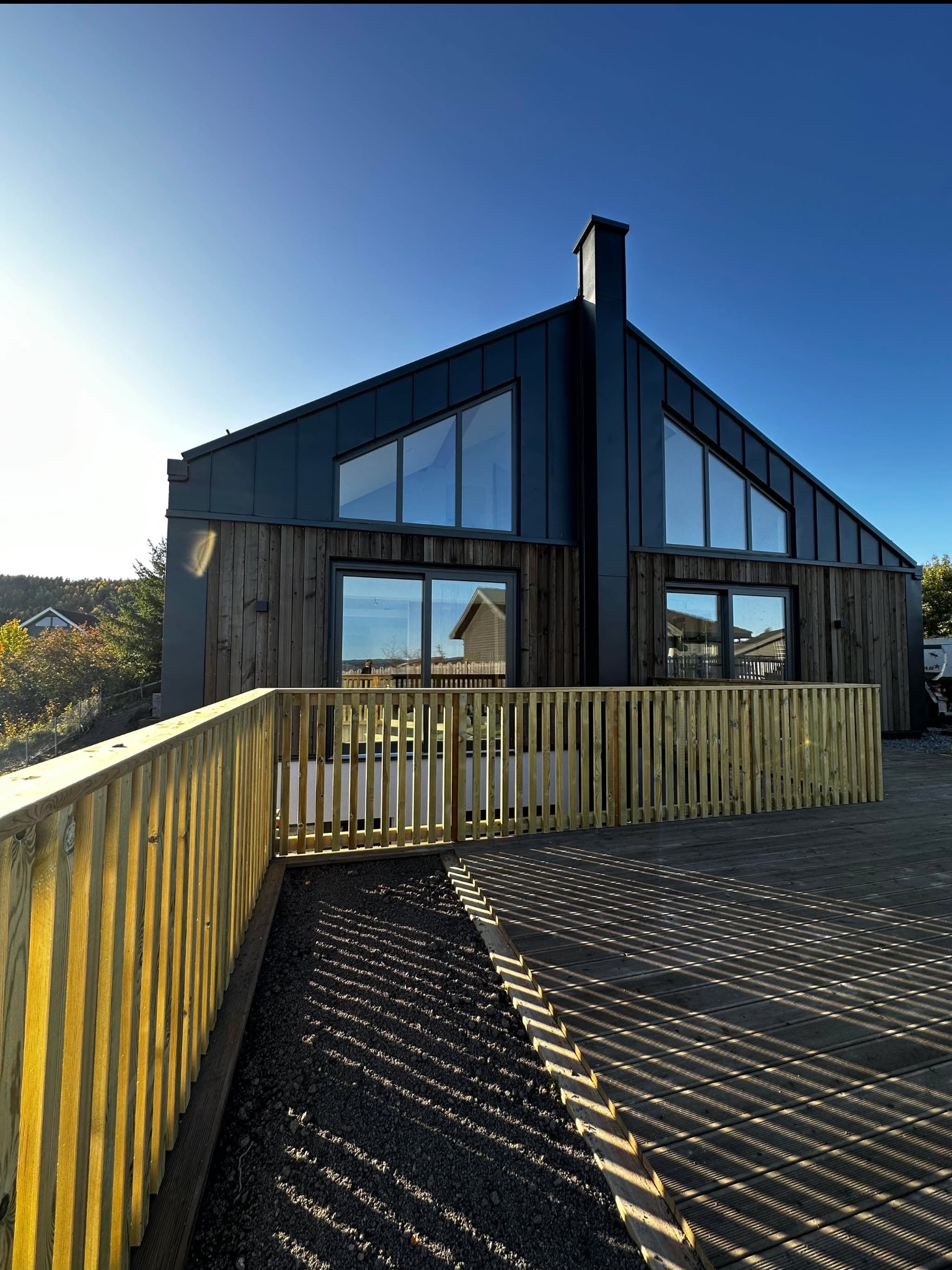 Nowoczesny dom szkieletowy z ciemną elewacją i dużymi oknami, otoczony drewnianym tarasem z ażurową balustradą w ciepłym odcieniu. Widok w słoneczny dzień.