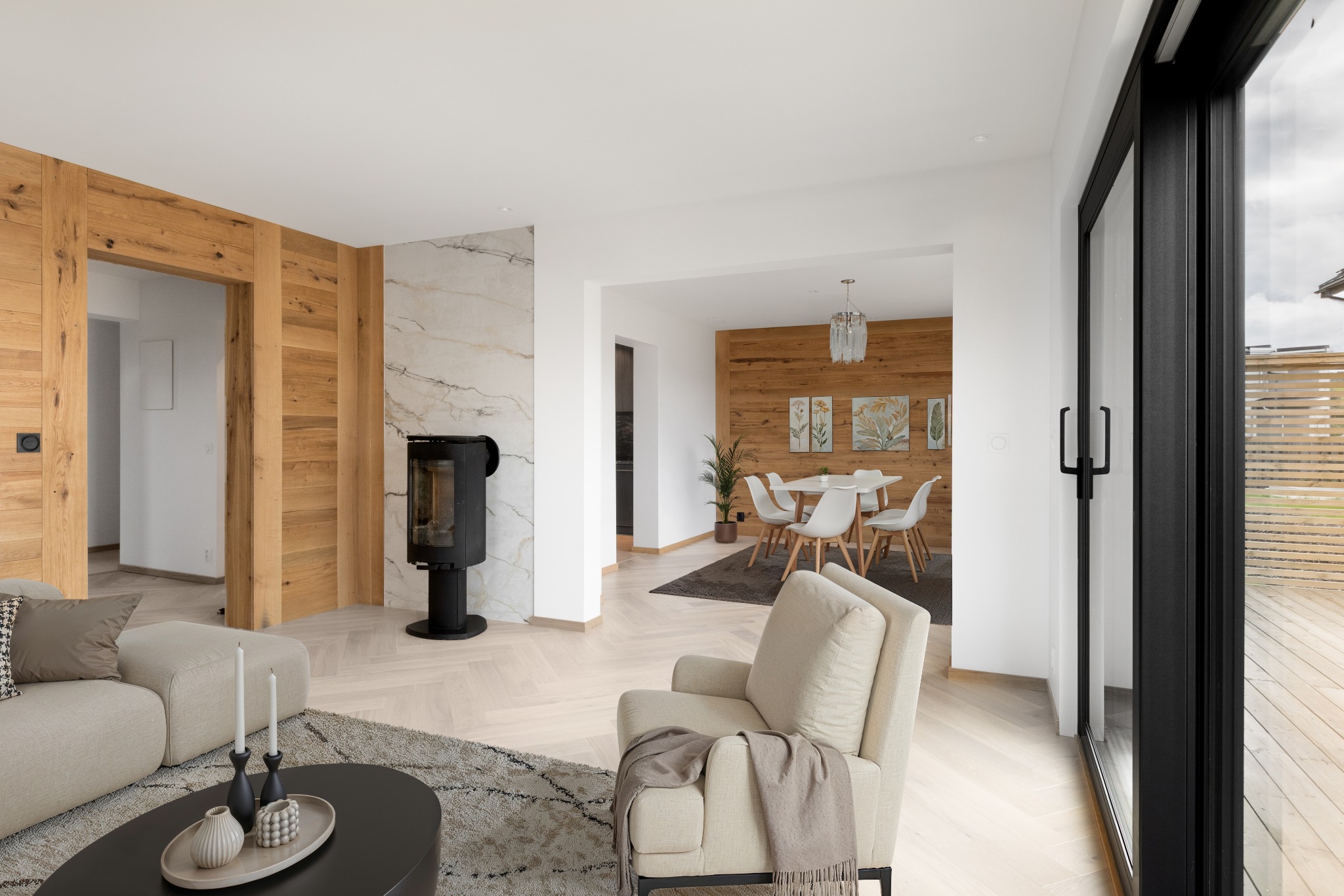 Jasne wnętrze z drewnianą ścianą i parkietem ułożonym w jodełkę. Widoczny kominek, sofa i krzesło. Duże okno z widokiem na zewnątrz doświetla przestrzeń.