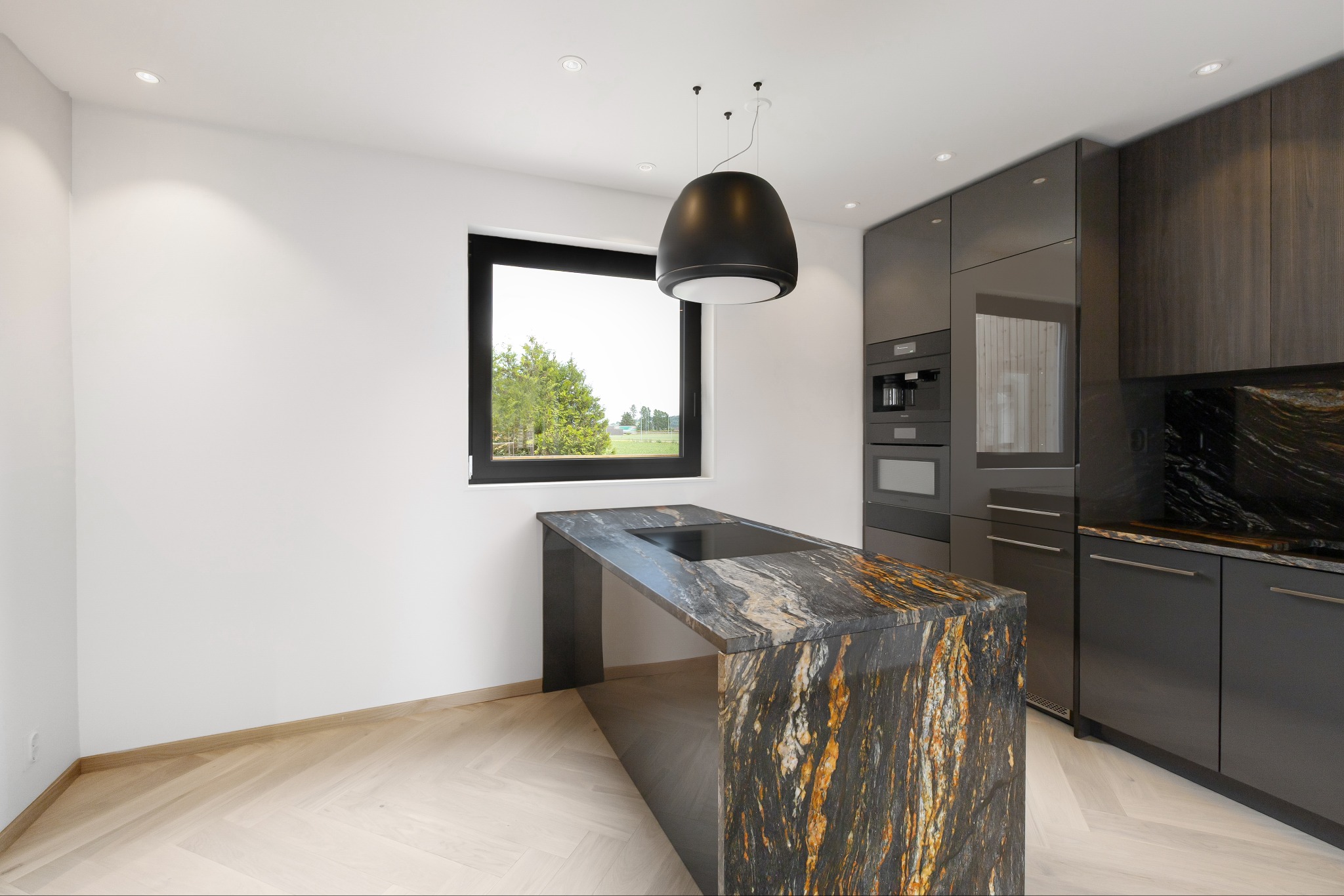 Nowoczesna kuchnia z wyspą z kamienia o unikalnym wzorze, drewnianą podłogą w jodełkę i minimalistycznym oświetleniem. Widok na zieleń przez okno.