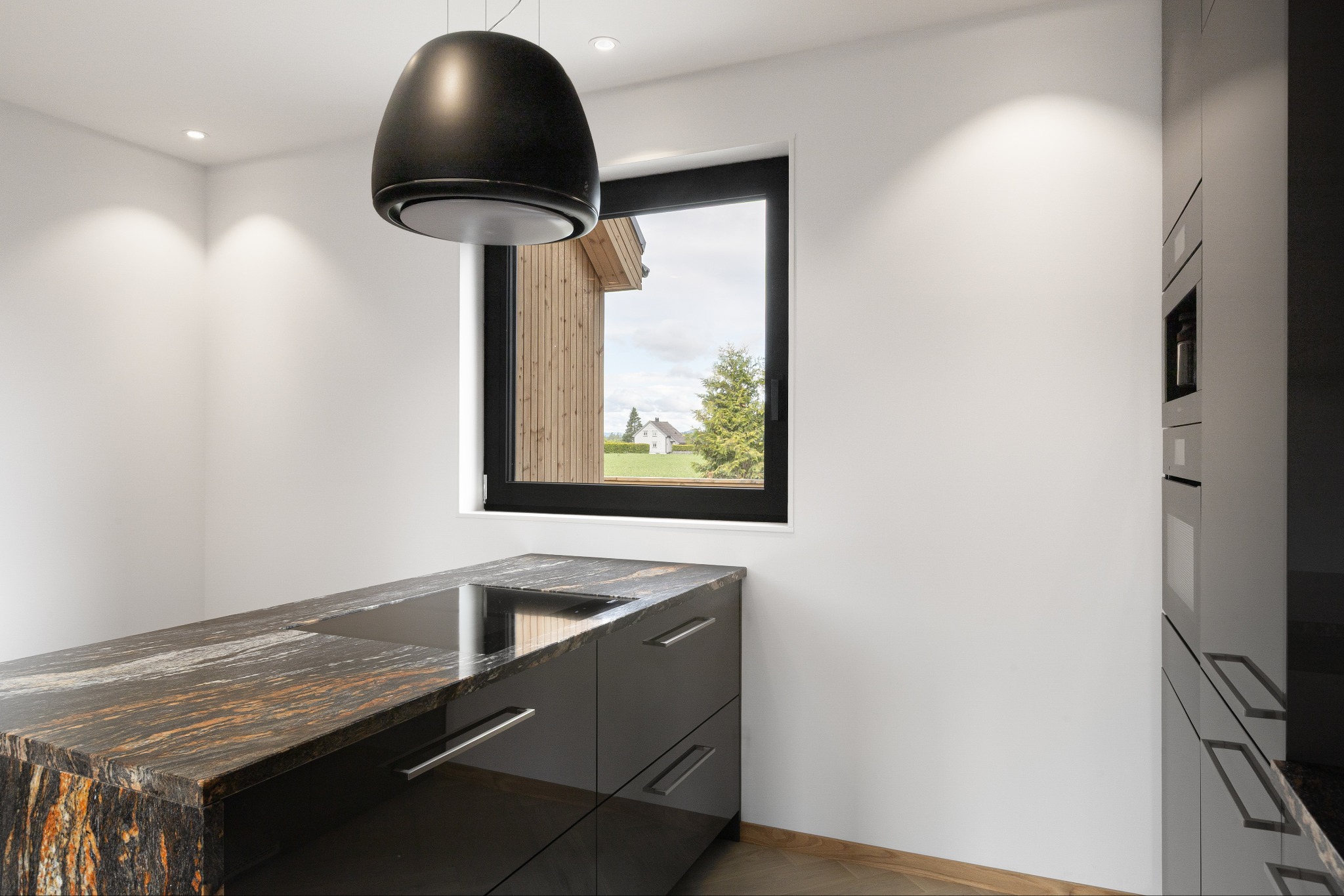 Nowoczesna kuchnia z blatem z kamienia, czarne szafki z metalowymi uchwytami, okno z widokiem na dom i ogród. Czarna lampa wisząca. Minimalistyczny design.