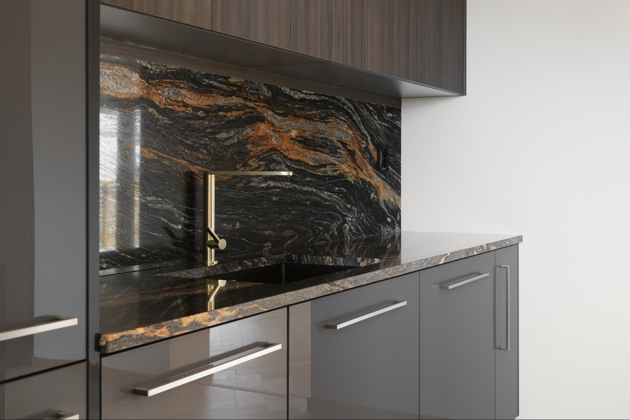 Błyszcząca, szara zabudowa kuchenna z blatem i panelem nad blatem z czarnego kamienia z pomarańczowymi żyłami. Złota bateria. Nowoczesny design kuchni.
