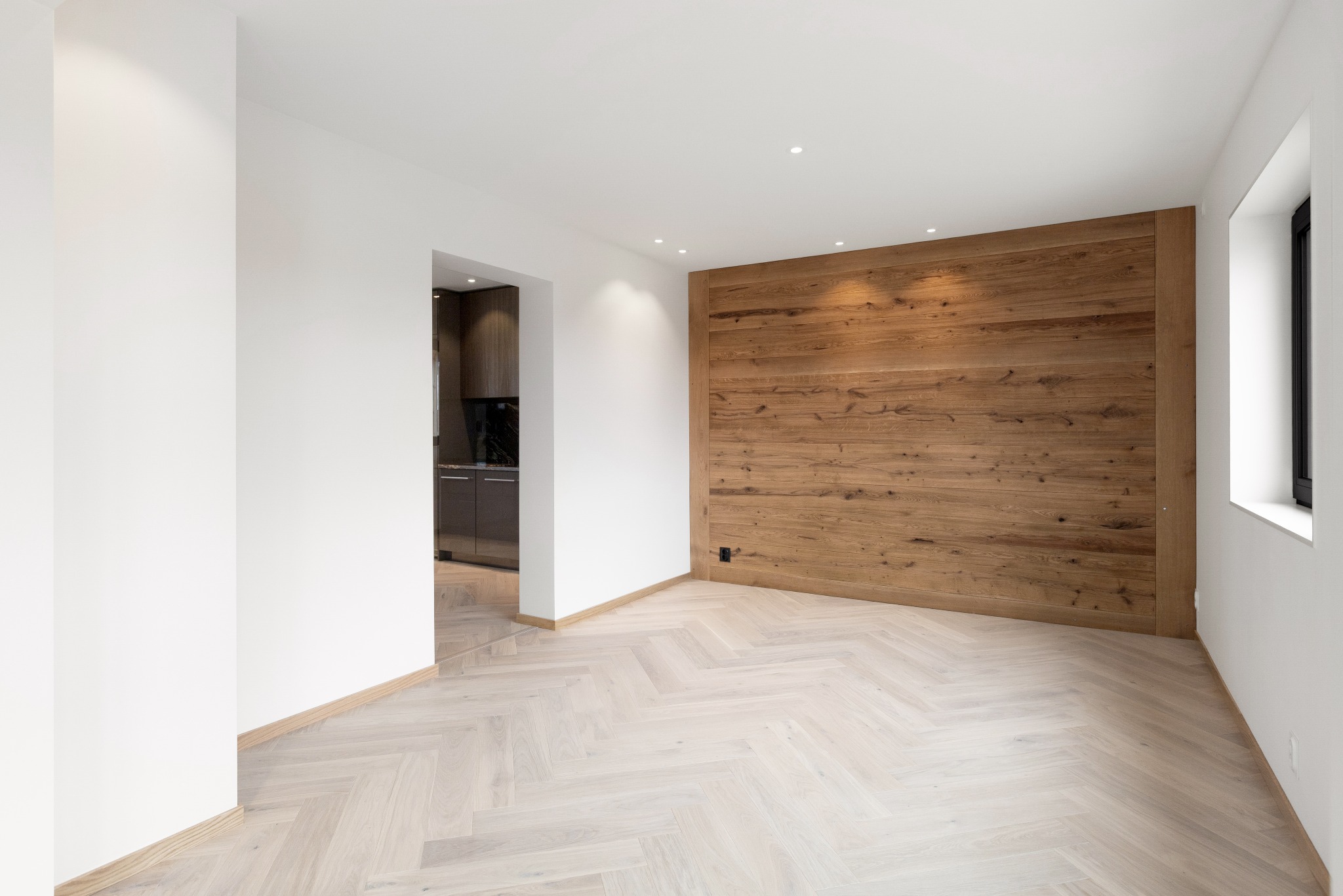 Jasny parkiet ułożony w jodełkę francuską w przestronnym wnętrzu z drewnianą ścianą akcentującą i widokiem na kuchnię. Minimalistyczny design.