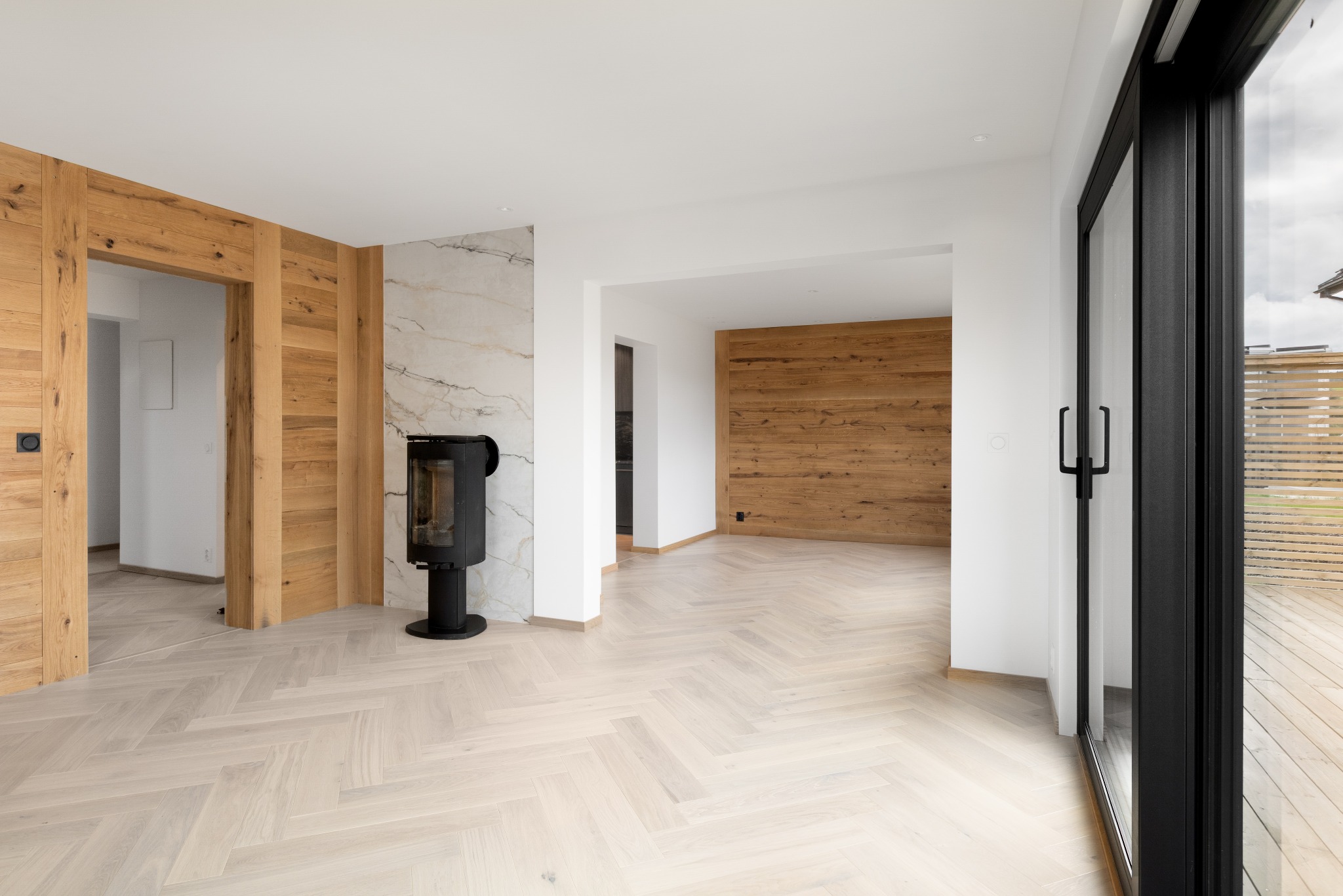 Jasna podłoga ułożona w jodełkę francuską, drewniane panele na ścianach i czarny piec w nowoczesnym wnętrzu z dużymi, przesuwnymi drzwiami balkonowymi.