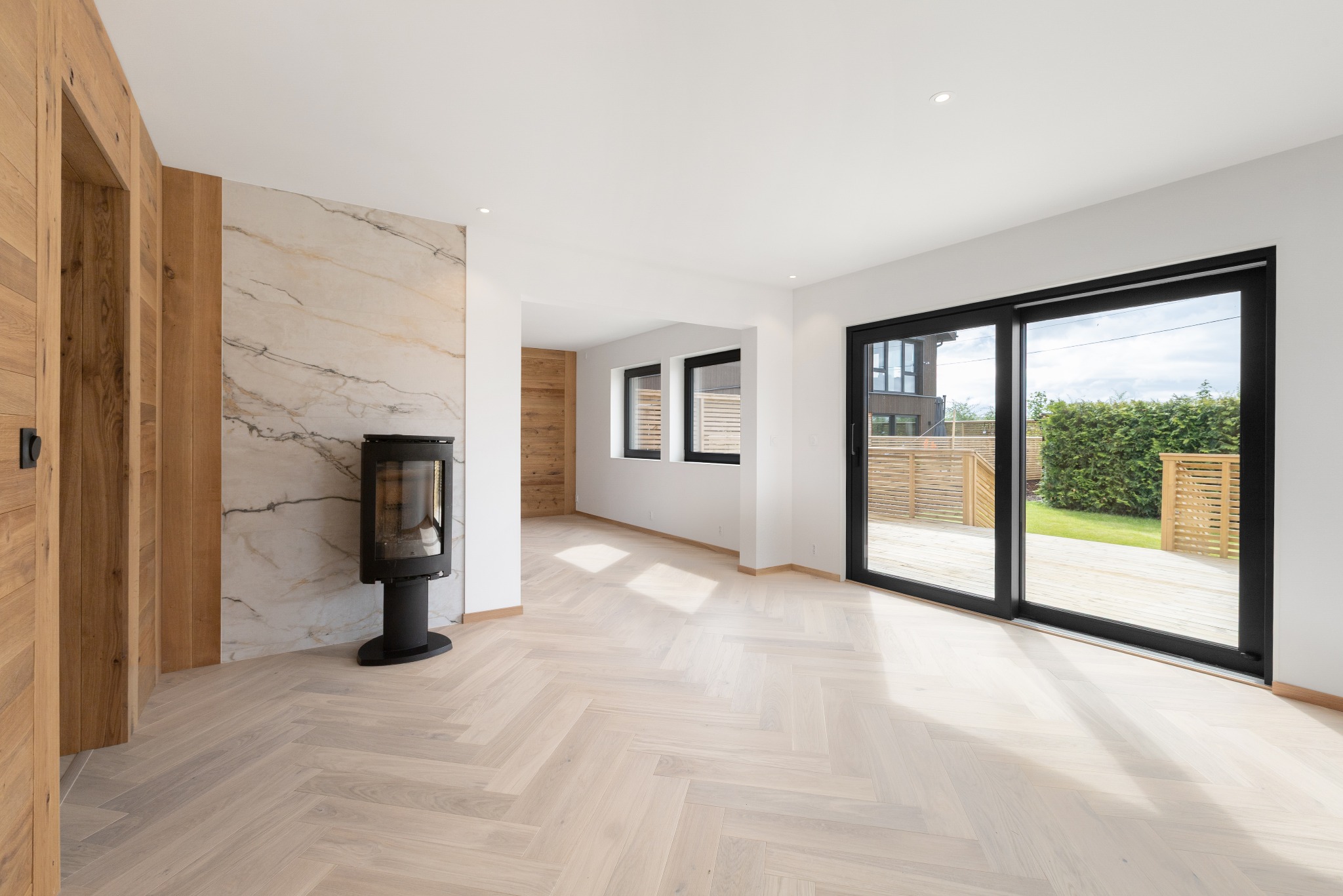 Jasna podłoga ułożona w jodełkę francuską w nowoczesnym salonie z kominkiem i dużymi przesuwnymi drzwiami balkonowymi z czarnymi ramami. Widok na taras i zieleń.