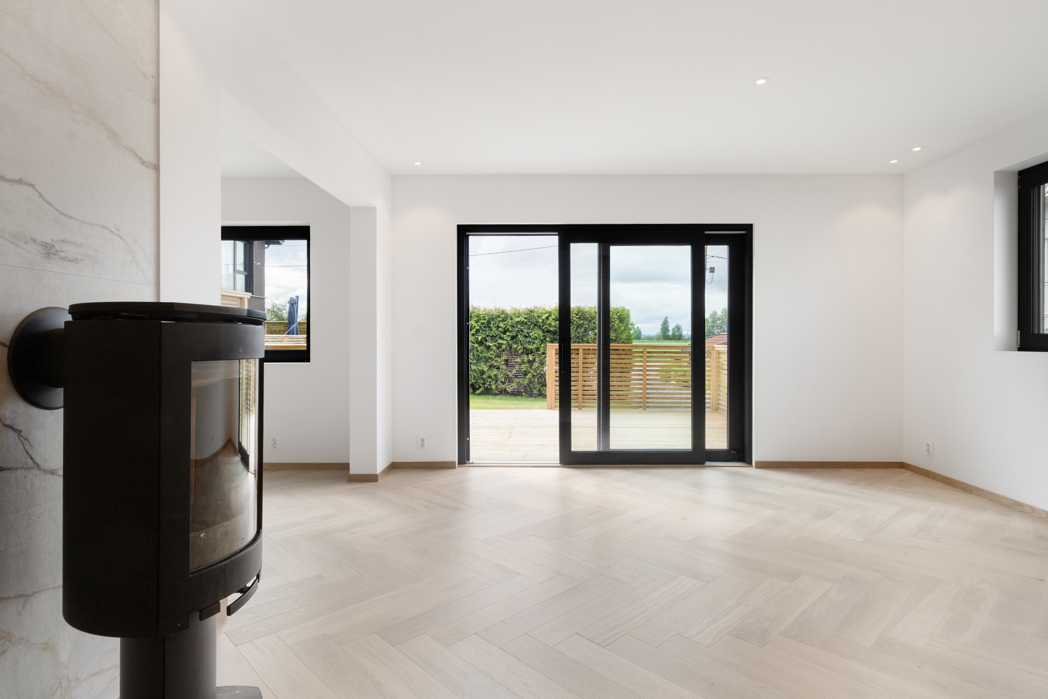 Jasny parkiet ułożony w jodełkę w nowoczesnym salonie z kominkiem i dużymi przesuwnymi drzwiami balkonowymi, widok na taras i zieleń. Minimalistyczne wykończenie.