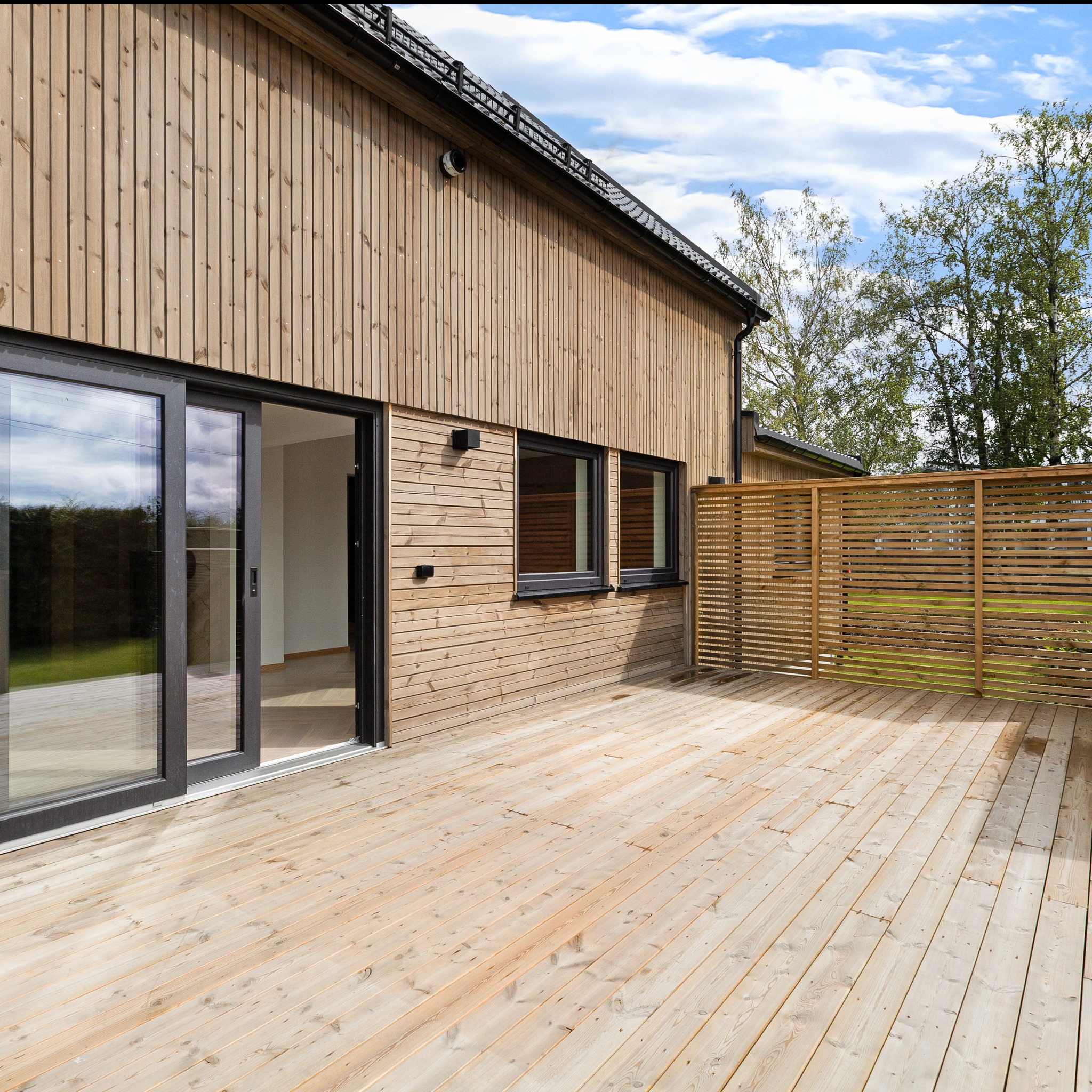 Drewniany dom szkieletowy z tarasem, jasne deski, przesuwne drzwi balkonowe, okna z czarnymi ramami, ażurowa drewniana przegroda, błękitne niebo w tle.