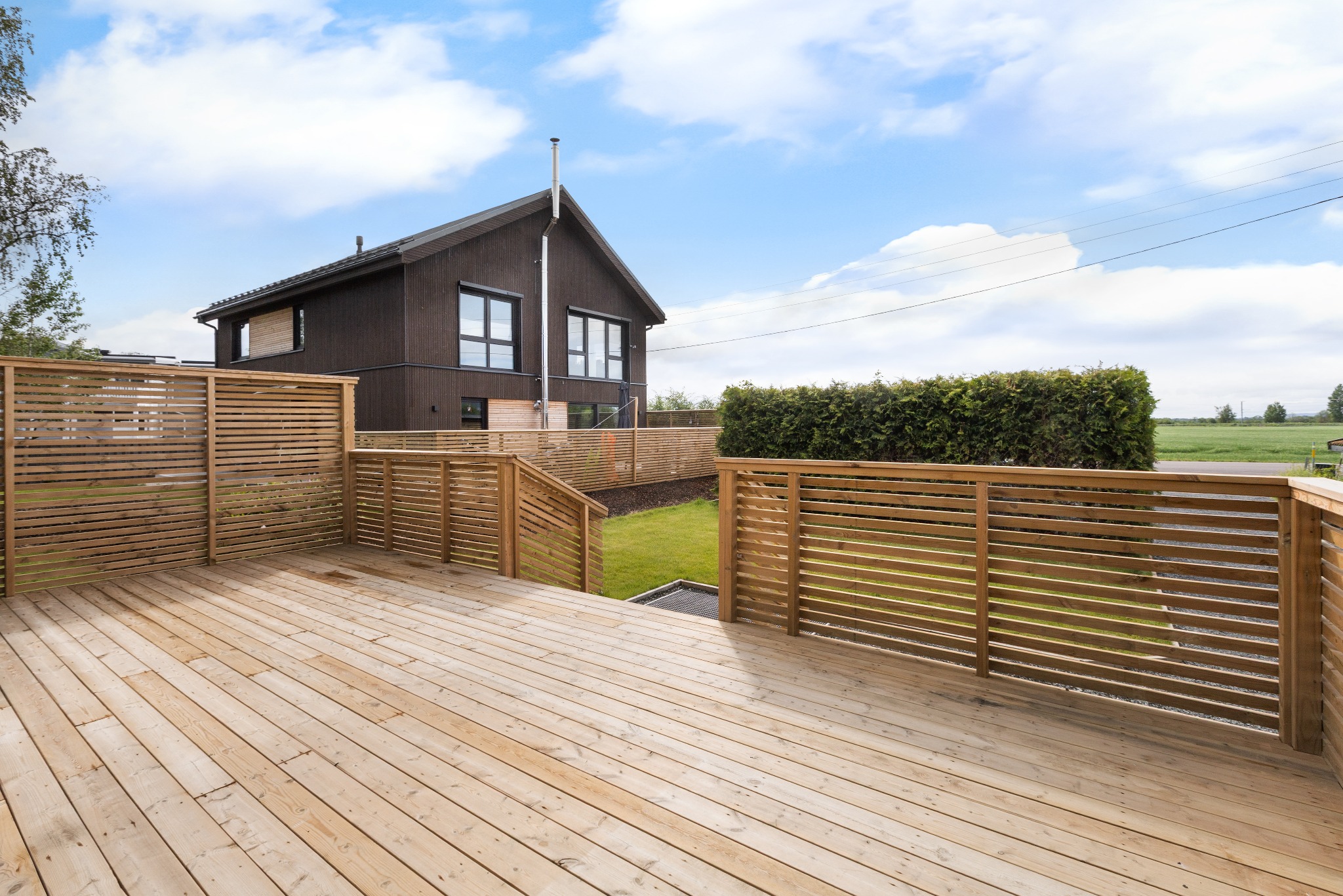 Drewniany taras z ażurowym ogrodzeniem przy nowoczesnym domu z ciemną elewacją. Widok na zadbany trawnik i błękitne niebo z chmurami.