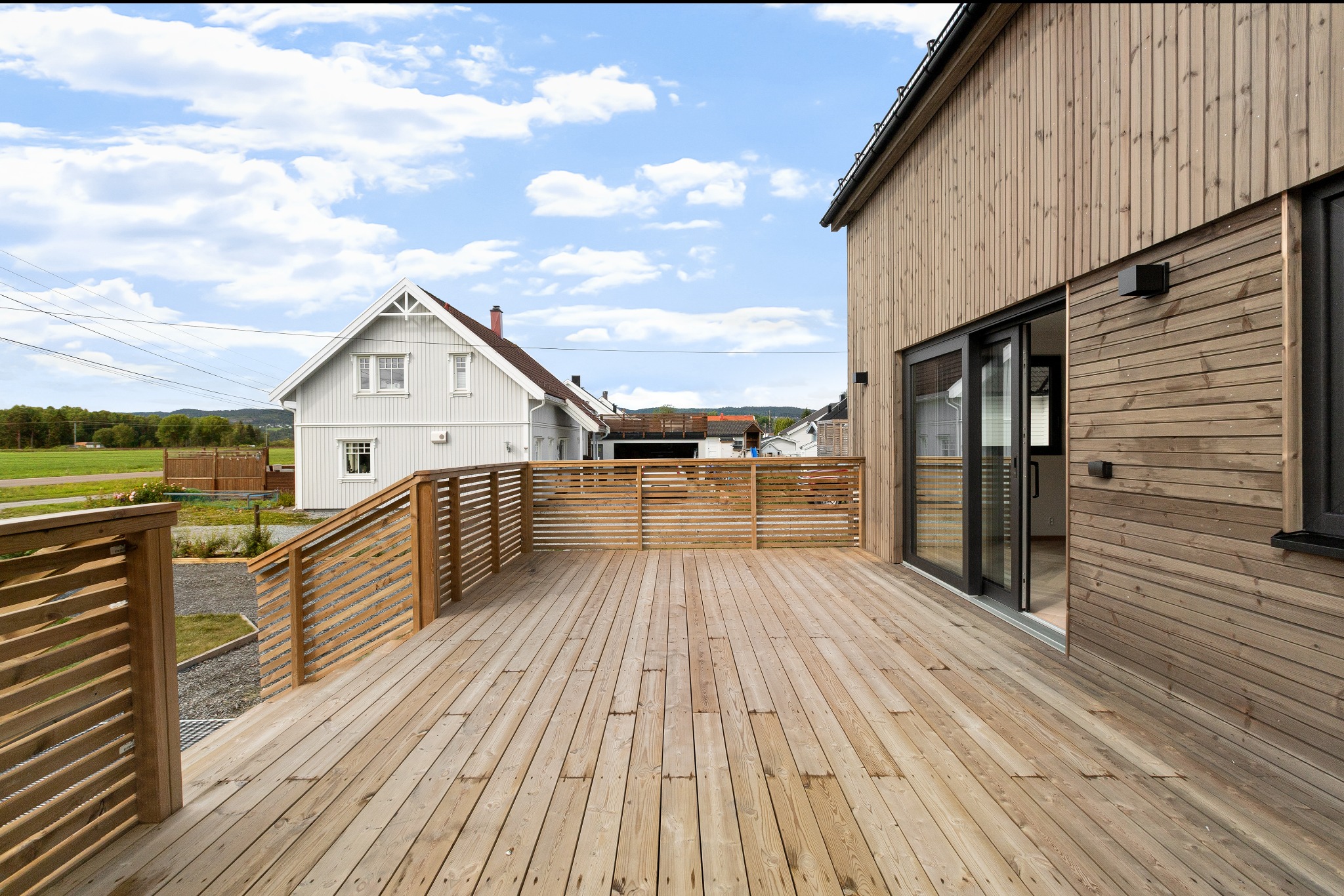 Drewniany taras z balustradą przy domu z drewnianą elewacją i przesuwnymi drzwiami. W tle dom w stylu skandynawskim i błękitne niebo.