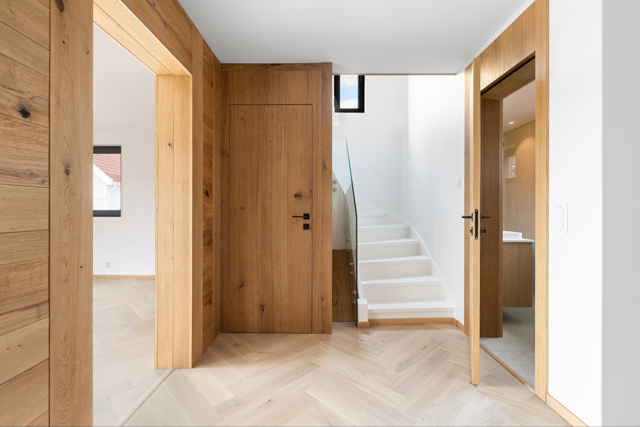 Nowoczesne wnętrze domu z drewnianymi drzwiami, panelami podłogowymi ułożonymi w jodełkę i białymi schodami ze szklaną balustradą. Minimalistyczny design.