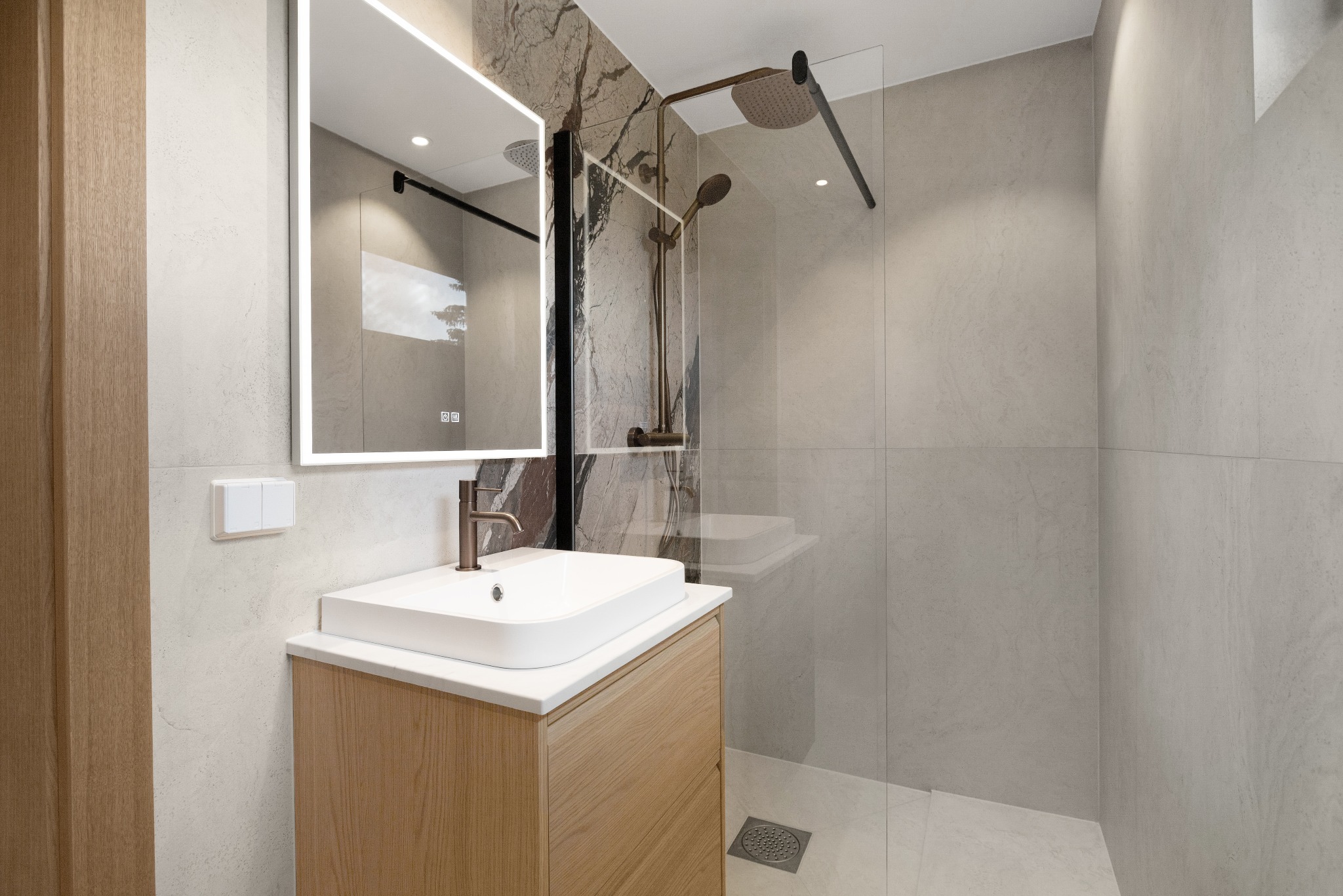 Nowoczesna łazienka z podświetlanym lustrem, umywalką na szafce z drewnianym fornirem i kabiną prysznicową z deszczownicą w kolorze brązu.