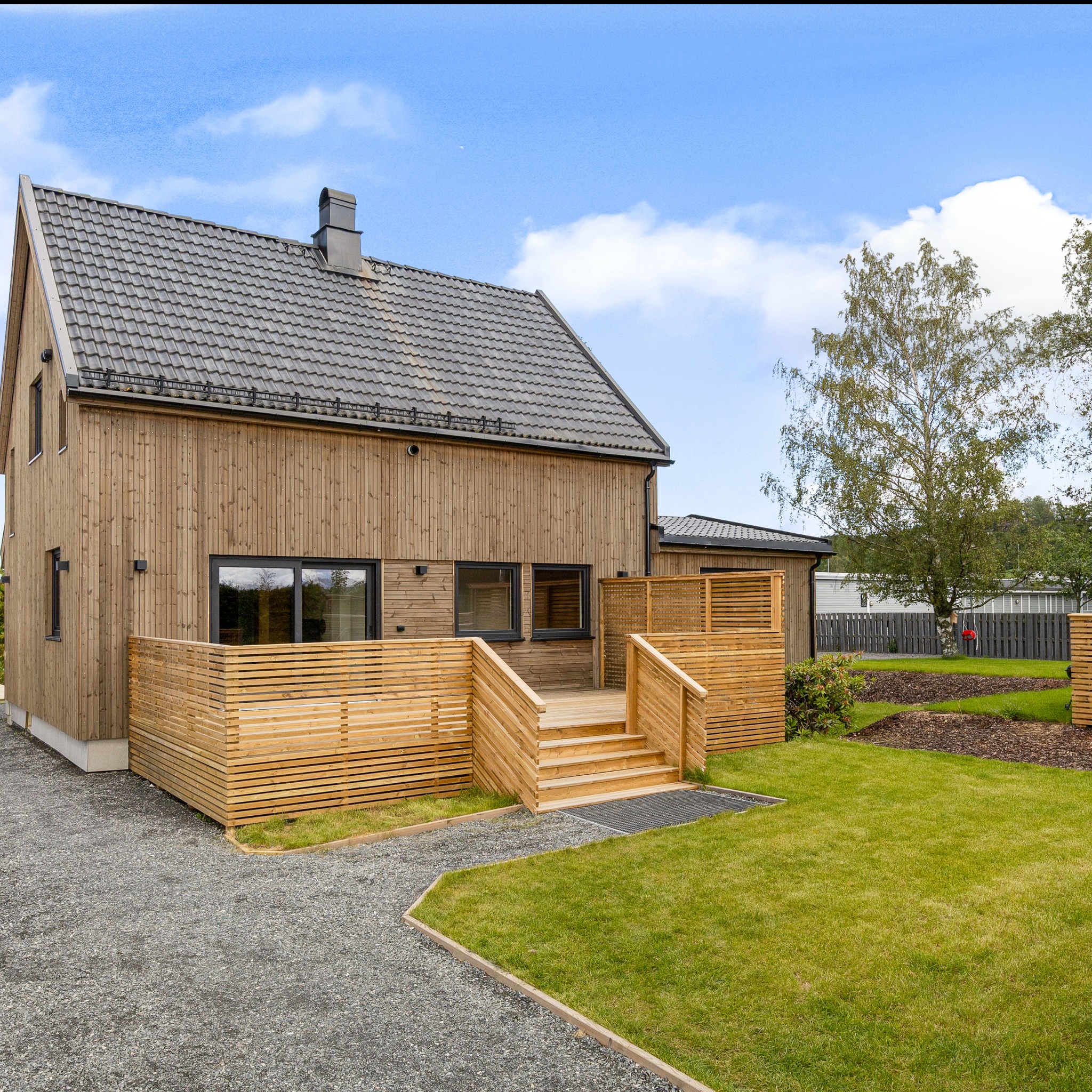 Nowoczesny dom szkieletowy z drewnianą elewacją i tarasem, otoczony zielonym trawnikiem i szarą nawierzchnią. Widoczne detale wykończenia i elementy architektury.